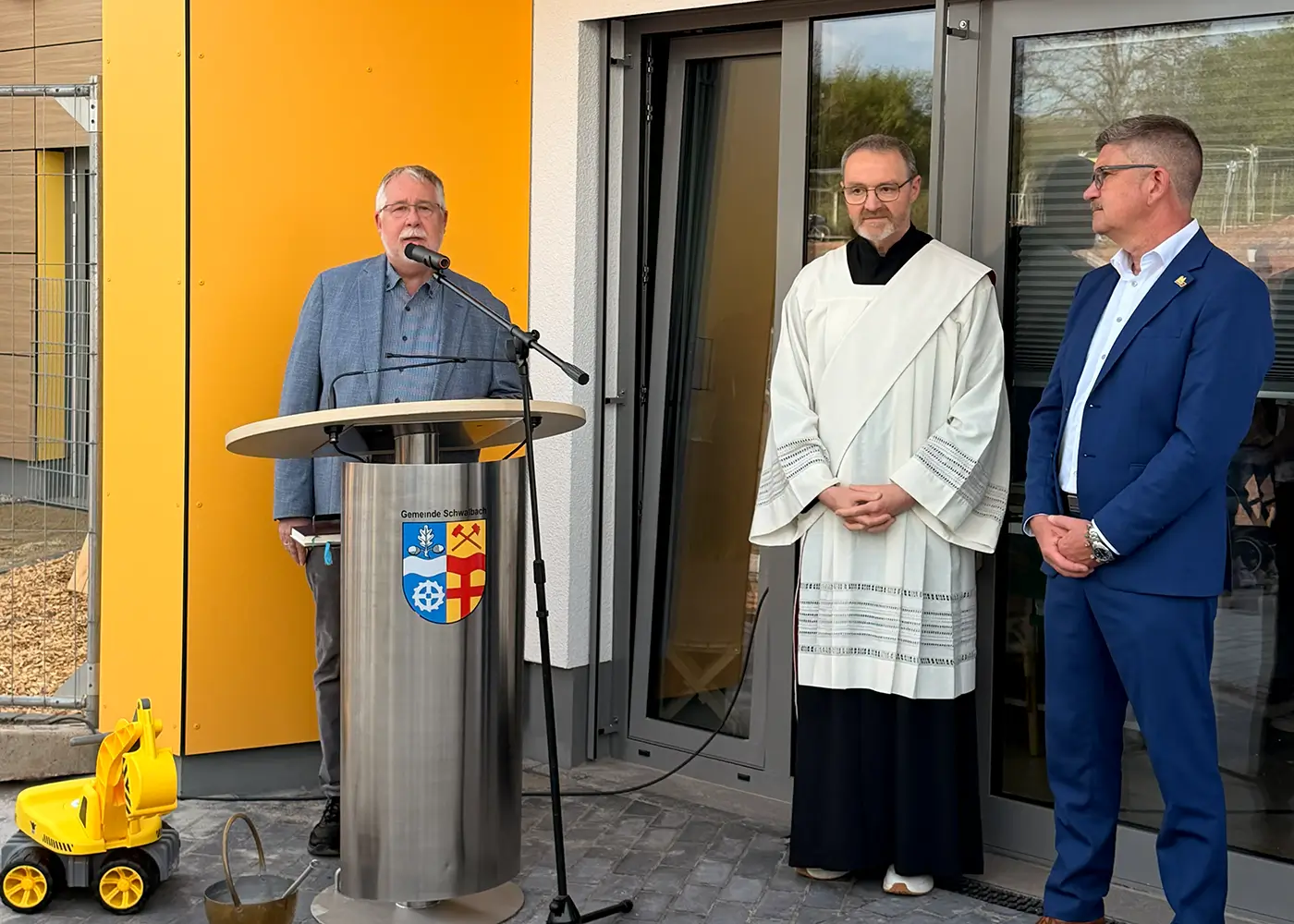 Drei Personen stehen vor einem Gebäude an einem Rednerpult der „Gemeinde Schmelzbach“.