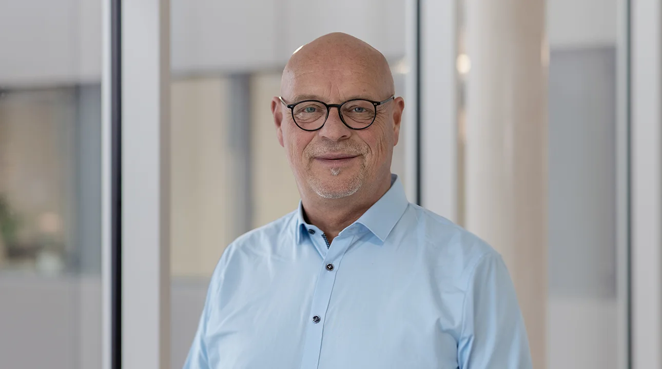 Porträt einer Person mit Brille und hellblauem Hemd in einem modernen Büro- oder Verwaltungsgebäude vor einer Fensterfront.