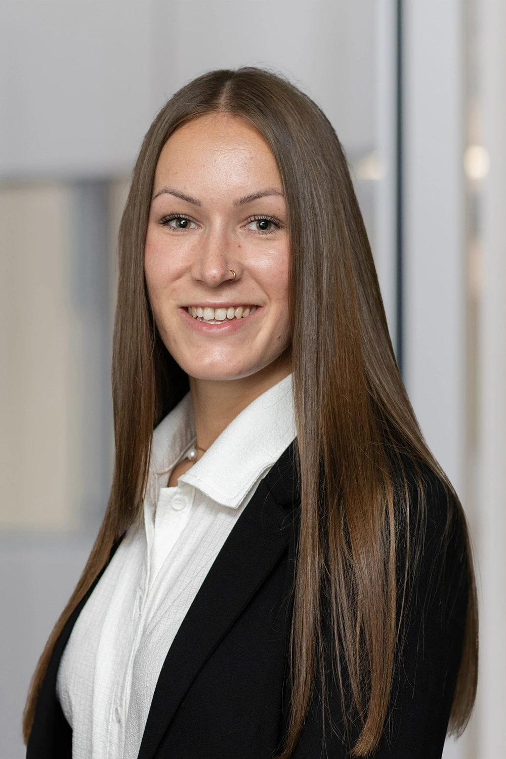 Porträt einer lächelnden Frau im schwarzen Blazer und weißer Bluse vor hellem Innenraumhintergrund