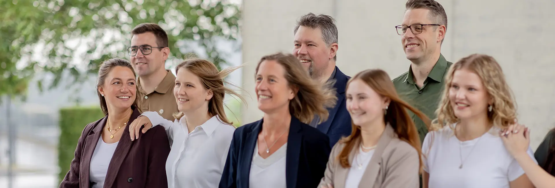 Gruppe von acht Personen in Business-Casual-Kleidung im Freien vor einem modernen Gebäude. Die Personen stehen dicht beieinander und blicken in dieselbe Richtung.