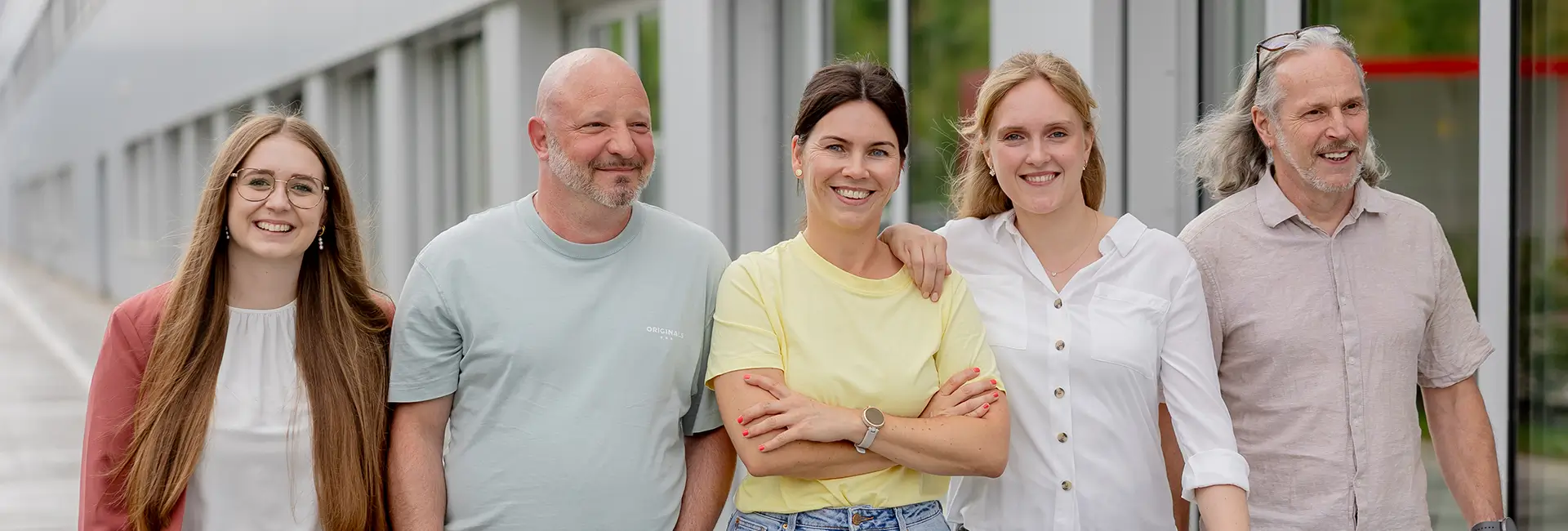 Fünf Personen stehen nebeneinander vor einer modernen Gebäudefassade mit großen Fenstern. Die Personen blicken in Richtung Kamera und tragen helle, legere Kleidung. Der Hintergrund zeigt einen überdachten Außenbereich mit Glasfronten.