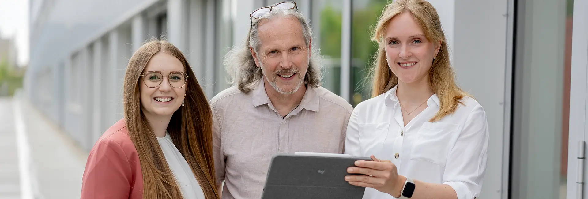 Drei Personen stehen vor einem modernen Bürogebäude und blicken in die Kamera. Eine Person hält ein Tablet, alle stehen eng beieinander in einem hellen Außenbereich mit großen Fensterfronten im Hintergrund.