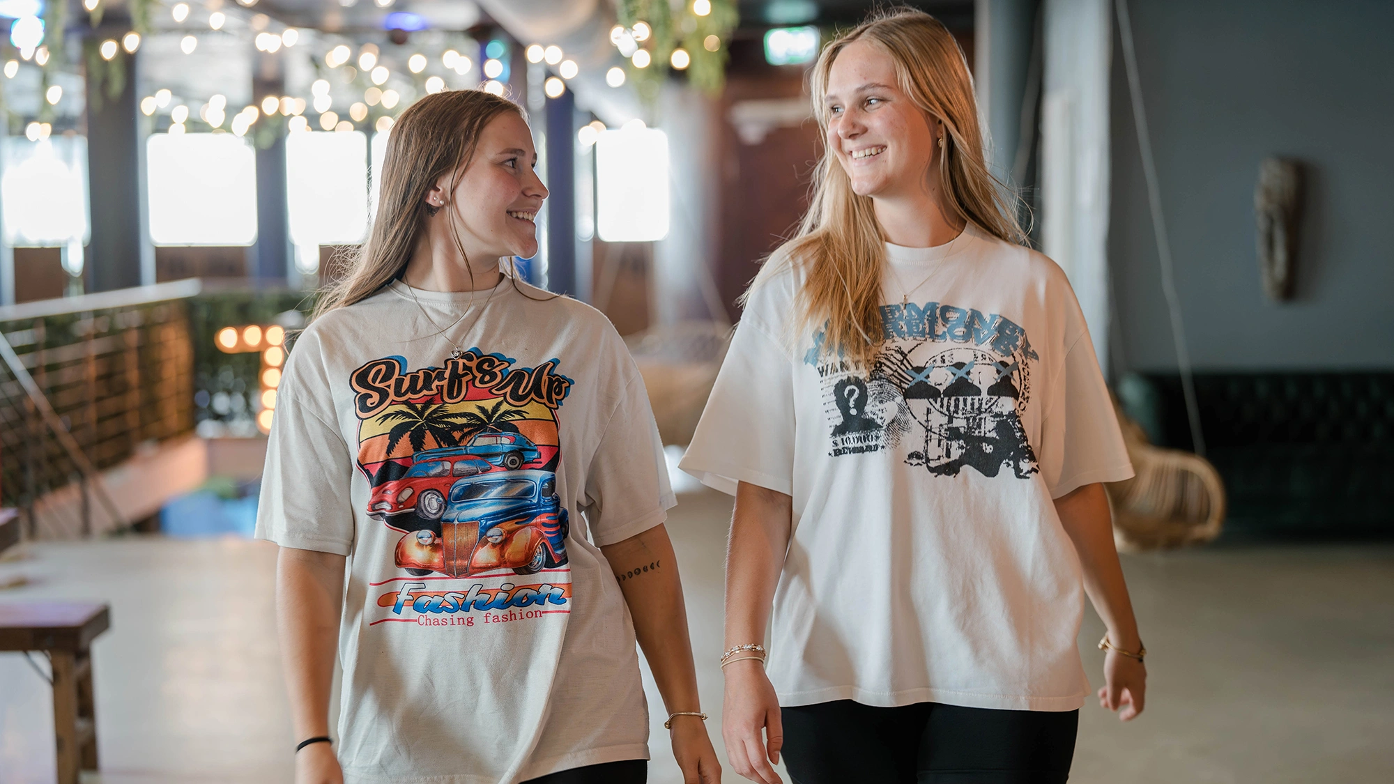 Zwei junge Frauen laufen lächelnd nebeneinander in einem hell erleuchteten Raum. Eine trägt ein T-Shirt mit der Aufschrift "Surf's Up Fashion Chasing fashion."
