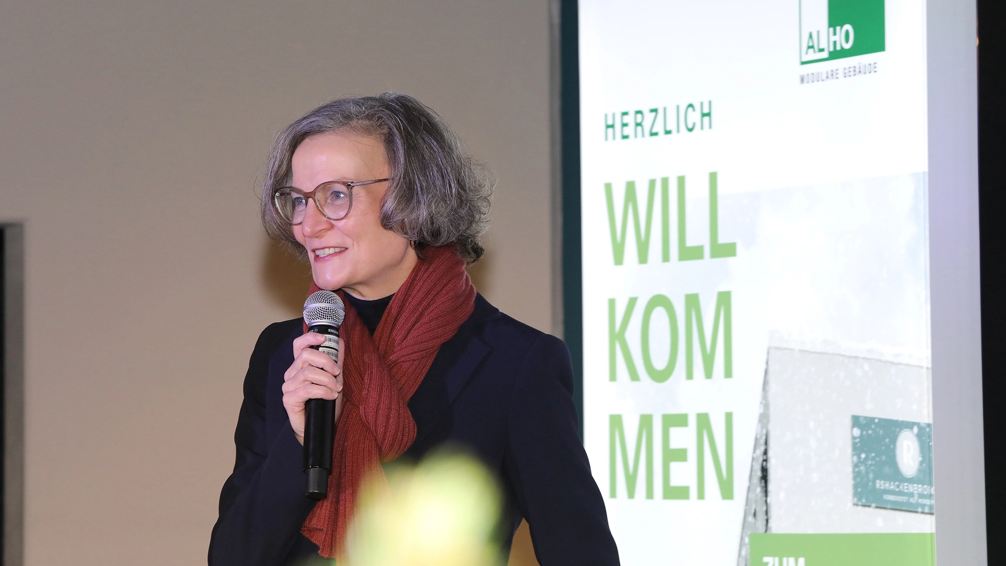 Frau hält bei einer Veranstaltung ein Mikrofon vor einer ALHO-Willkommenspräsentation