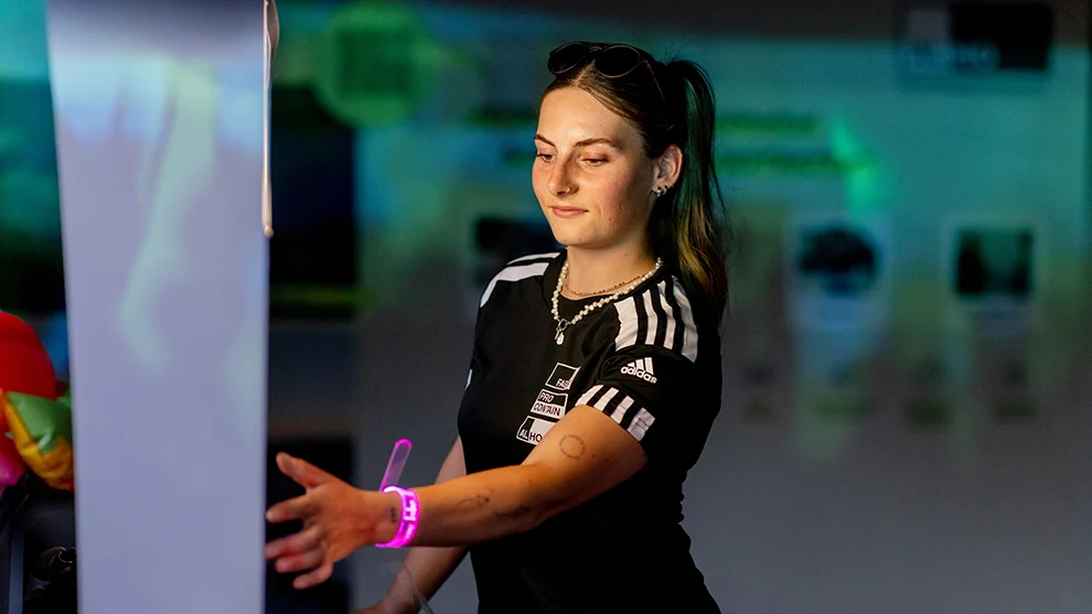 Eine Frau trägt ein schwarzes Trikot mit "Pro Contain" und Adidas-Logo, bewegt den Arm nach vorne in einem beleuchteten Raum, während sie ein leuchtendes Armband trägt.