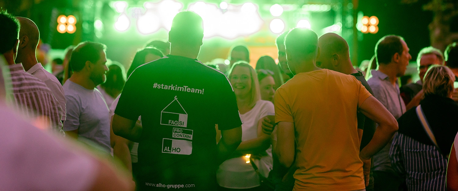 Person mit blauem T-Shirt mit der Aufschrift „#starkimTeam! FAGSI PRO CONTAIN ALHO www.alho-gruppe.com“ steht in Menschenmenge bei grün beleuchtetem Event.