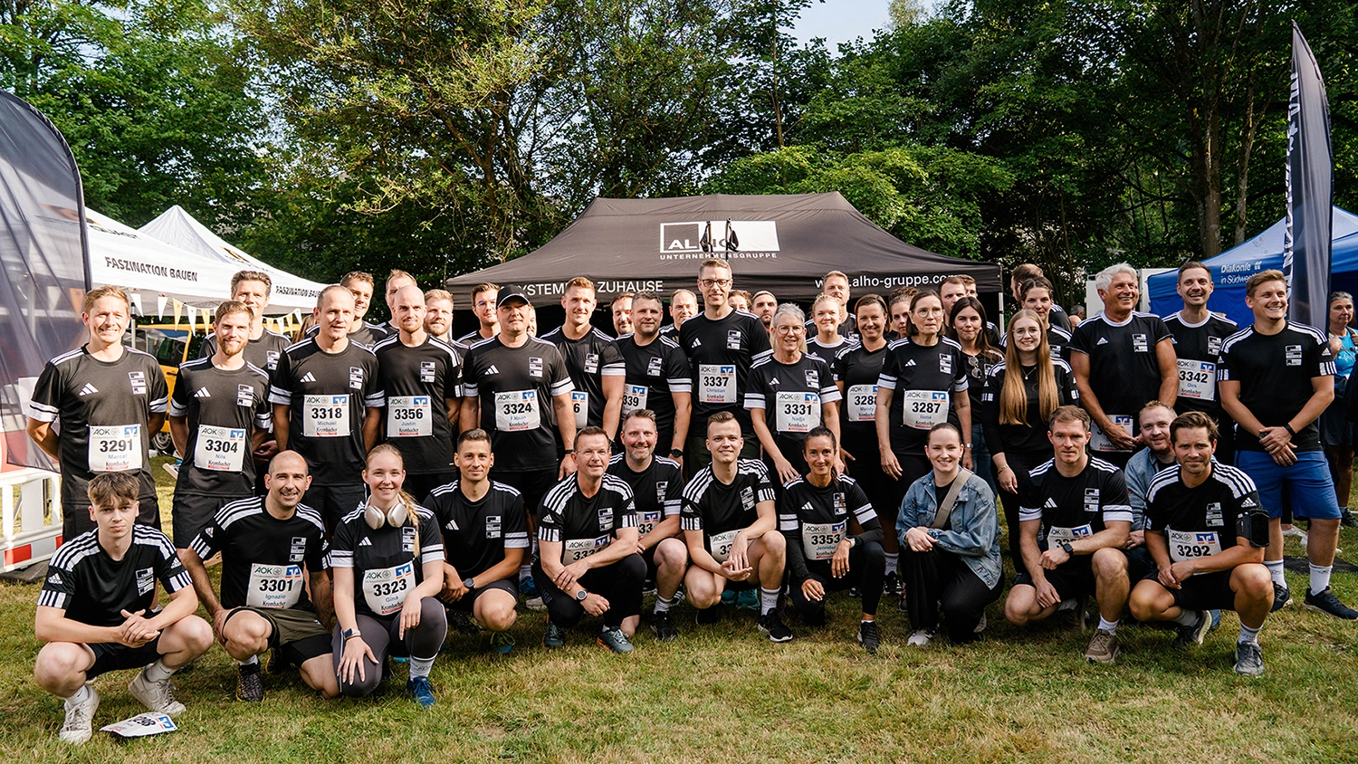 Gruppenfoto von Mitarbeitenden der ALHO Unternehmensgruppe in Sportkleidung mit Startnummern bei einer Laufveranstaltung, aufgenommen vor einem ALHO-Eventzelt im Grünen