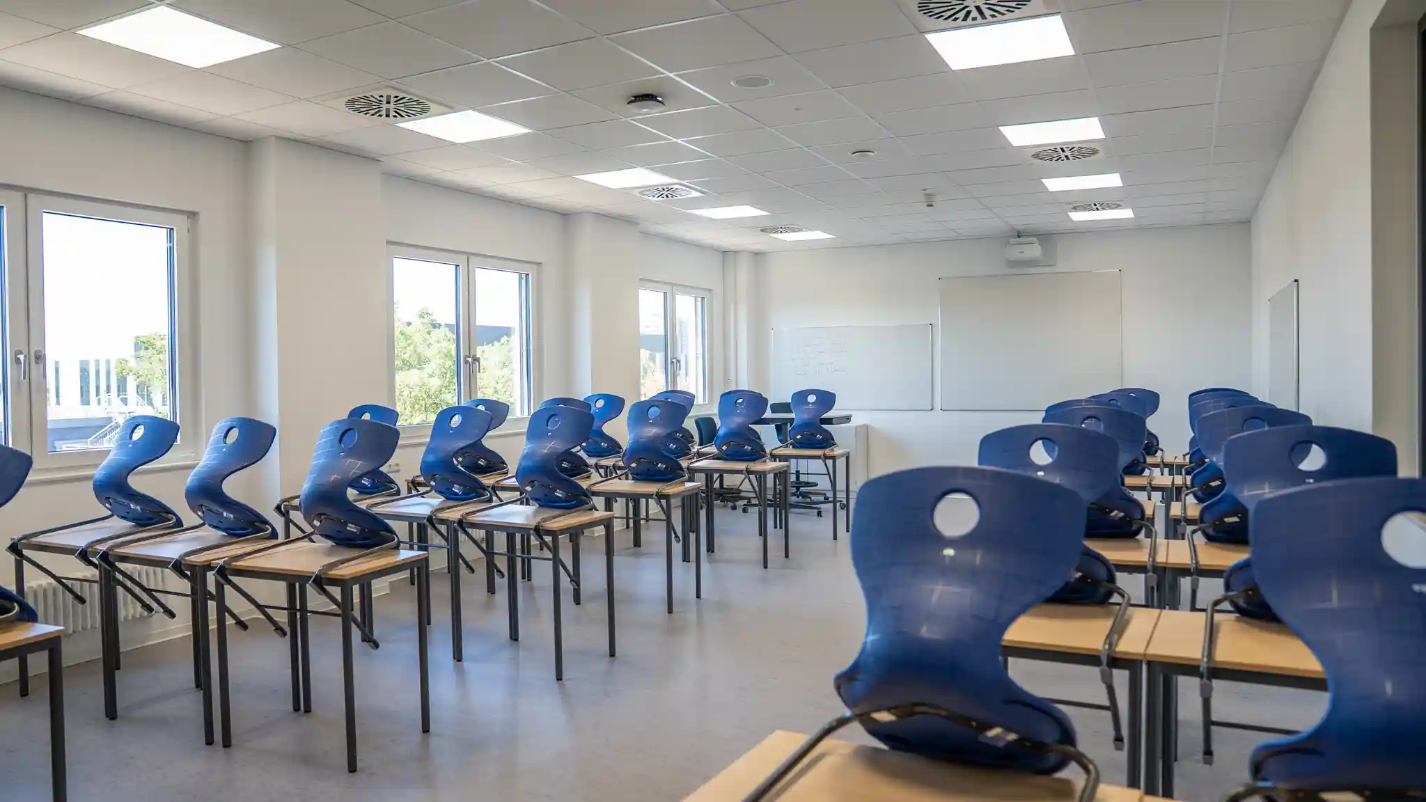 Ein heller, moderner Klassenraum mit großen Fenstern, weißen Wänden und mehreren Reihen von Schülertischen. Die blauen Stühle stehen auf den Tischen, der Raum ist ungenutzt und für den Unterricht vorbereitet. Im vorderen Bereich befinden sich Whiteboards und Präsentationsflächen.