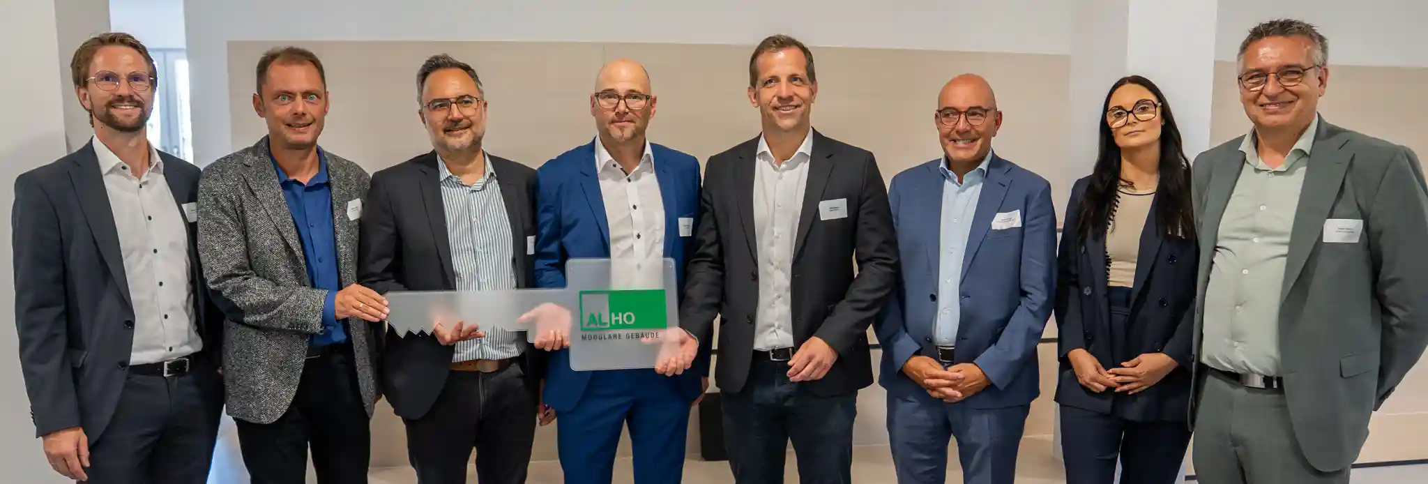 Gruppenfoto mit mehreren Vertreterinnen und Vertretern des Bauherrn, der Schule und der ALHO Systembau GmbH im neuen Schulgebäude. In der Mitte wird ein symbolischer großer Schlüssel mit ALHO-Logo präsentiert, der die Übergabe des Erweiterungsbaus der IGS Europa Mainz markiert. Die Aufnahme entstand in einem hellen Innenraum vor einer neutralen Wand.
