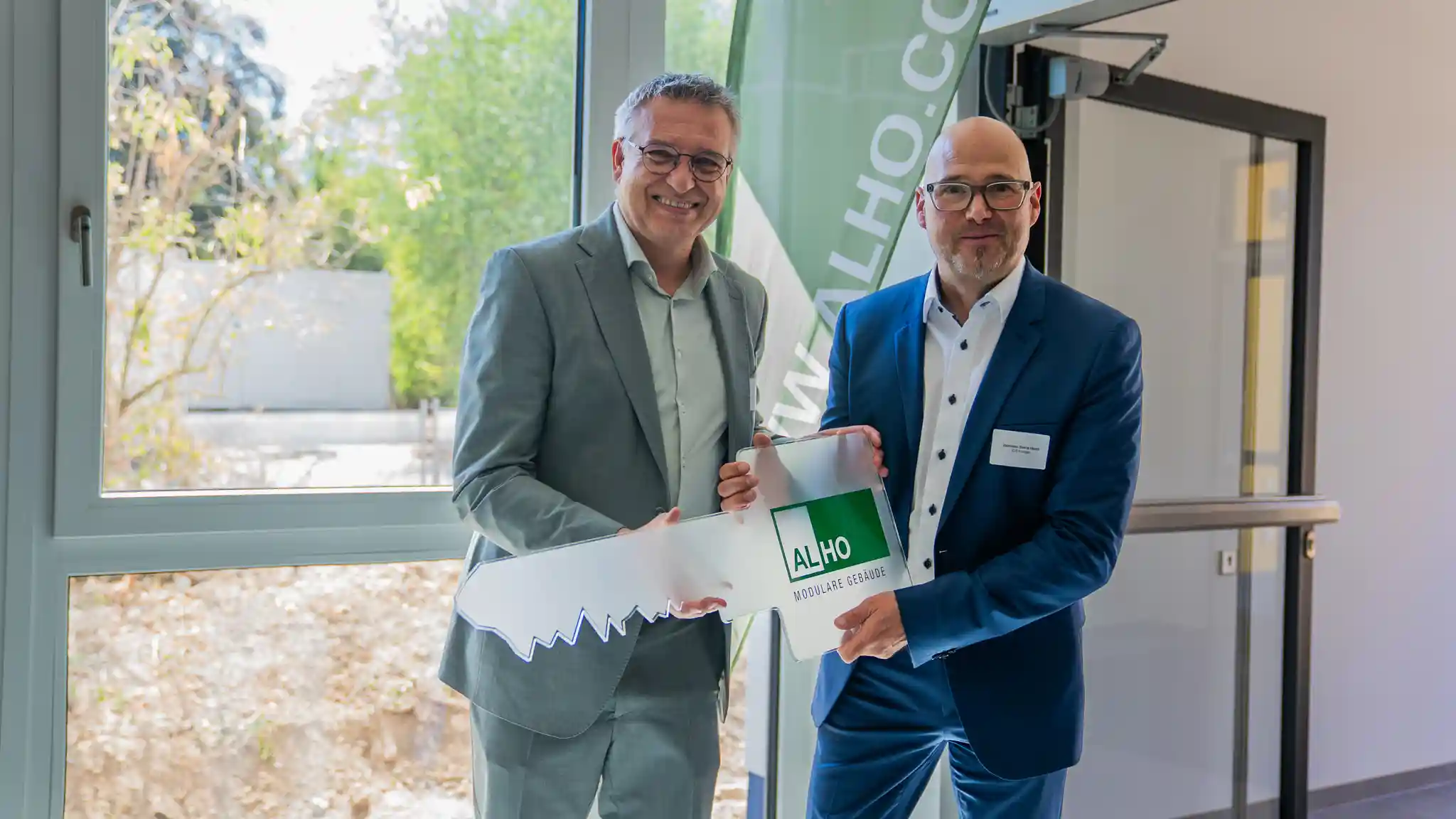 Zwei Personen in Business-Kleidung stehen in einem hellen Innenraum vor einer großen Fensterfront und halten gemeinsam eine ALHO-Präsentationsmappe mit dem Schriftzug „ALHO Modulare Gebäude“. Im Hintergrund ist ein grünes Roll-up-Banner mit teilweise sichtbarem ALHO-Logo zu sehen. Tageslicht fällt durch die Fenster und beleuchtet die Szene.