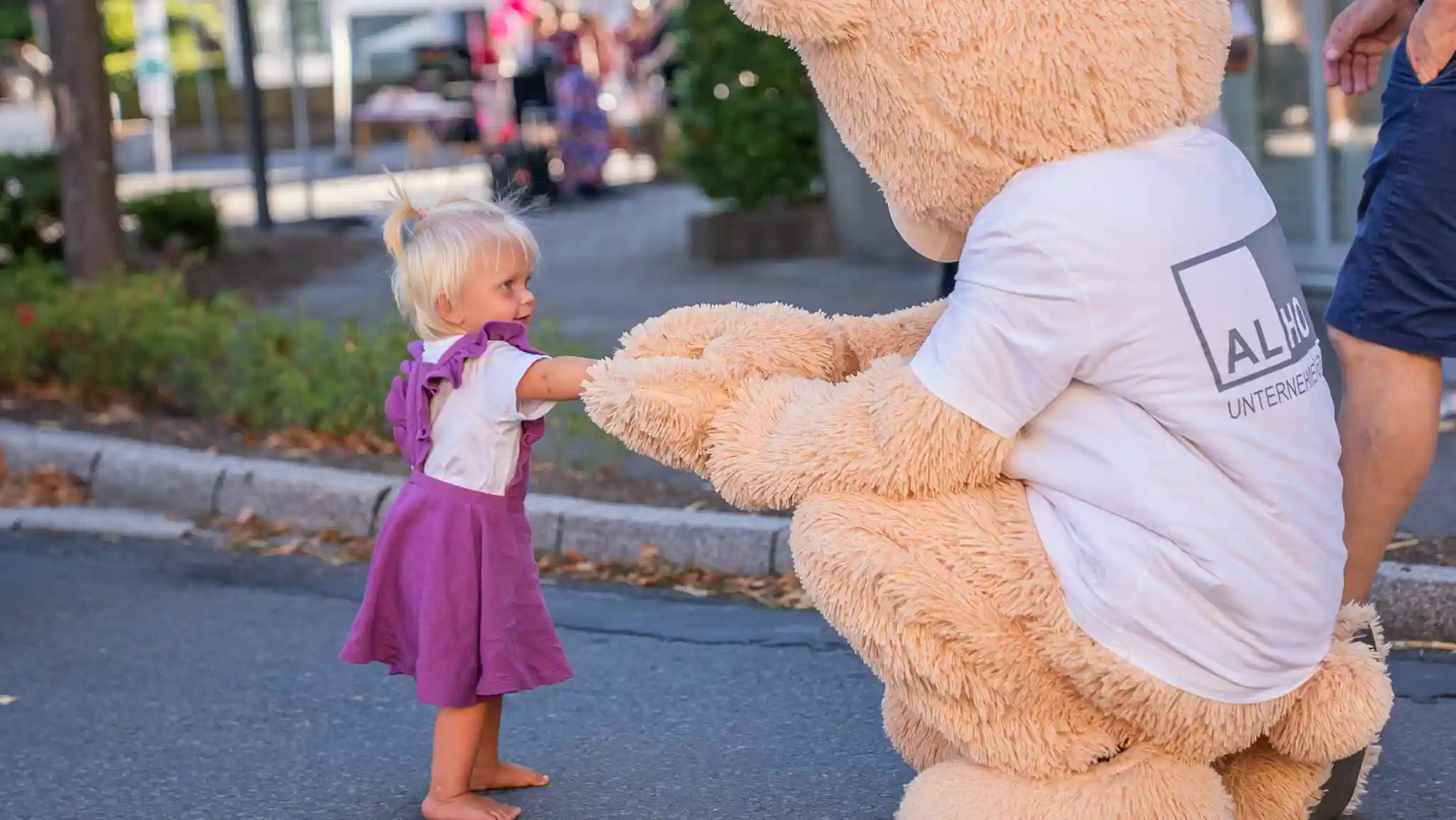 Ein kleines Kind in einem lila Kleid steht barfuß auf einer Straße und berührt die Hand eines großen Bärenmaskottchens mit ALHO-Logo bei einer Veranstaltung im Freien.