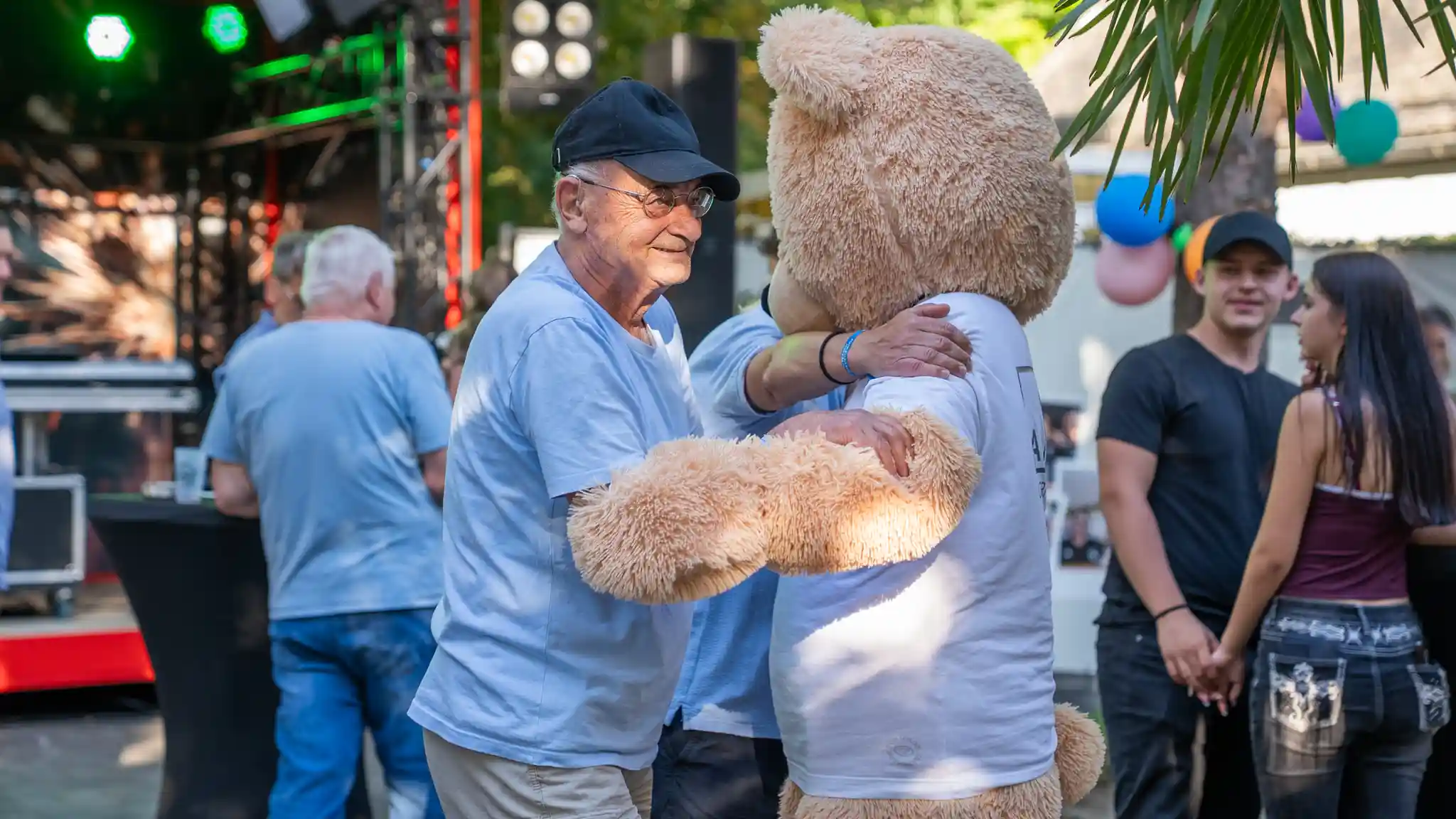 Eine Person in heller Kleidung begrüßt ein großes Bärenmaskottchen bei einer Veranstaltung im Freien. Im Hintergrund sind eine Bühne, Dekorationen und weitere Besucherinnen und Besucher zu sehen.