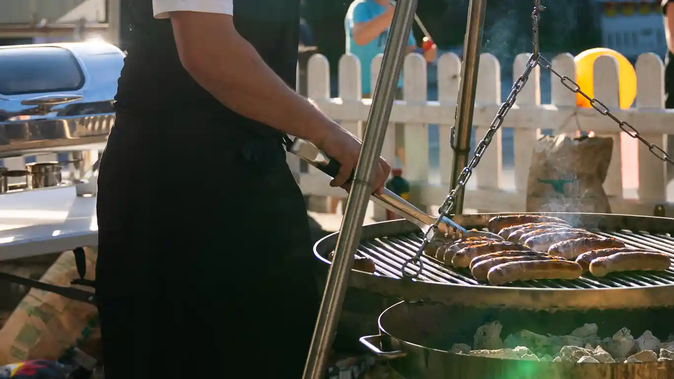 Eine Person wendet mehrere Würstchen auf einem runden Holzkohlegrill. Rauch steigt auf, während im Hintergrund eine Veranstaltungsfläche mit Zaun und Besuchenden zu sehen ist.