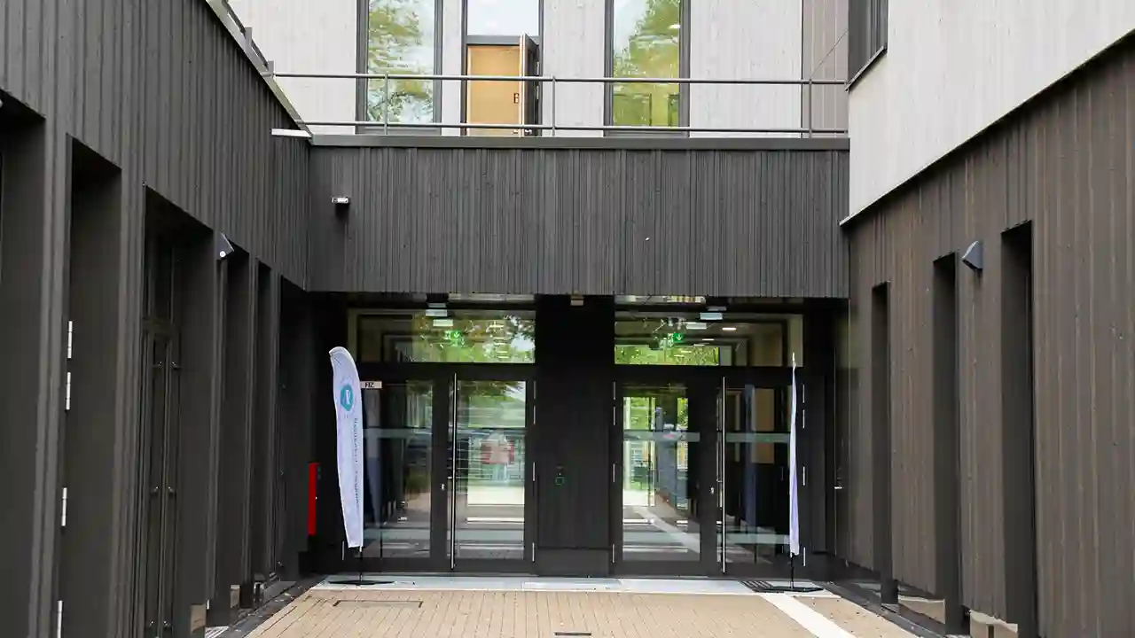 Moderner Eingangsbereich eines mehrgeschossigen Schul- oder Verwaltungsgebäudes mit dunkler Fassadenverkleidung, verglasten Eingangstüren und offenem Innenhof.